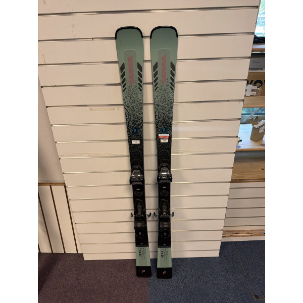 K2 Disruption SC W ski's volledig overzicht vooraanzicht met mintgroene en zwarte graphics en Marker ER3 10 GripWalk bindingen, hangend aan een slatwall in een skiwinkel