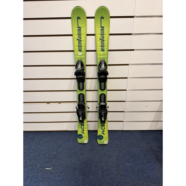 Paire complète de skis enfant Elan ACE Jett 100 verts avec lettrage Elan bleu et fixations, devant un mur à lamelles