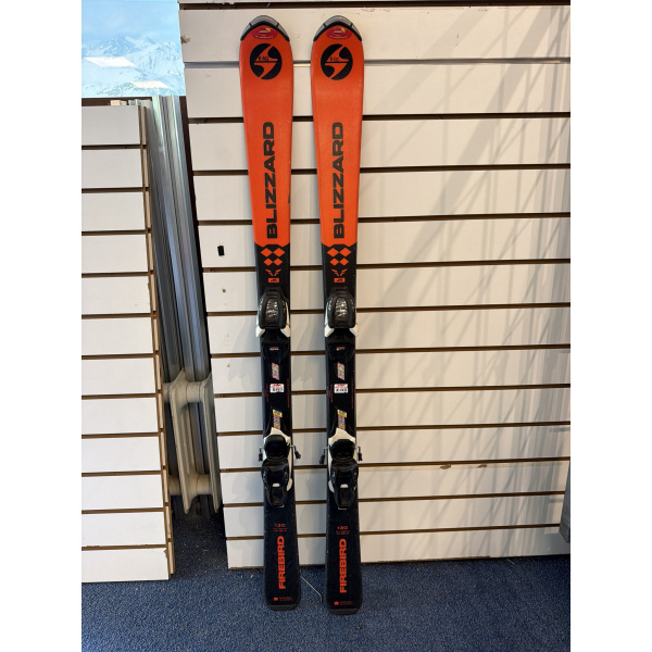 Paire complète de skis junior Blizzard Firebird JR aux spatules orange, moitié basse noire, avec fixations, devant un mur à lamelles