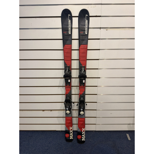 Paire complète de skis junior Elan Maxx Red noirs et rouges, longueur 140, fixations noires, devant un mur à lamelles