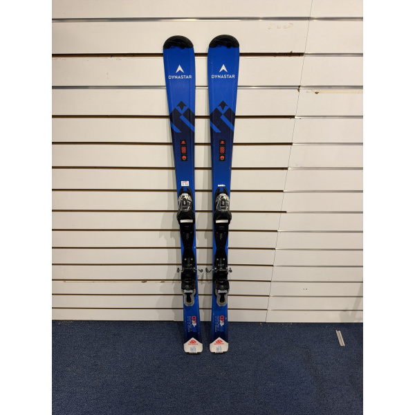 Paire complète de skis junior Dynastar Team Speed bleus avec logo Dynastar blanc et fixations Look, devant un mur à lamelles