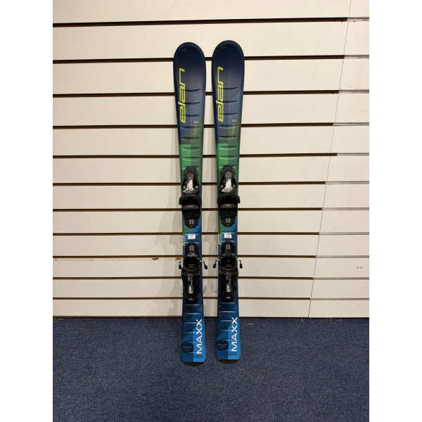 Paire complète de skis enfant Elan Maxx Blue à spatules bleu marine, partie basse verte et bleue, avec fixations, devant un mur à lamelles