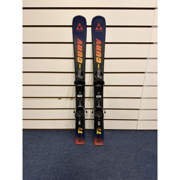 Paire complète de skis enfant Fischer The Curv Jr. bleu foncé avec logo Fischer rouge et inscription jaune, devant un mur à lamelles