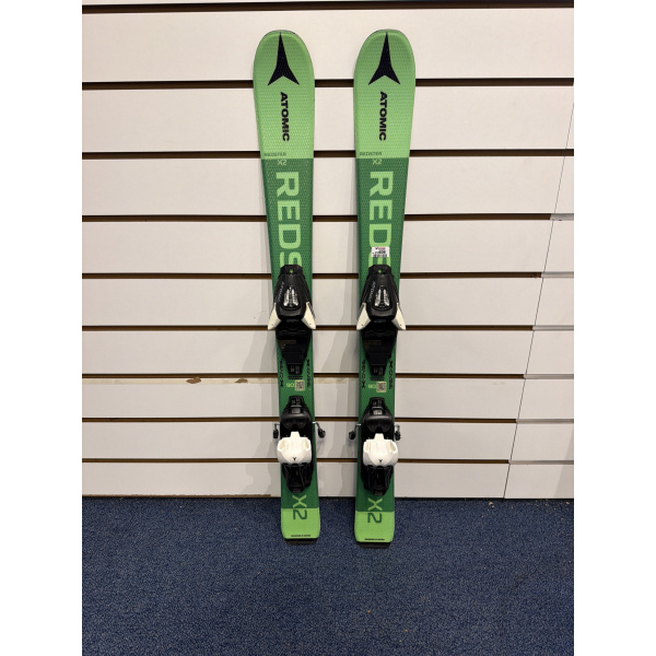 Paire complète de skis enfant Atomic Redster X2 verts avec lettrage ATOMIC noir et fixations, devant un mur à lamelles