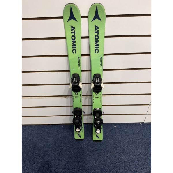Paire complète de skis enfant Atomic Redster X2 vert vif avec lettrage ATOMIC bleu foncé et fixations, devant un mur à lamelles