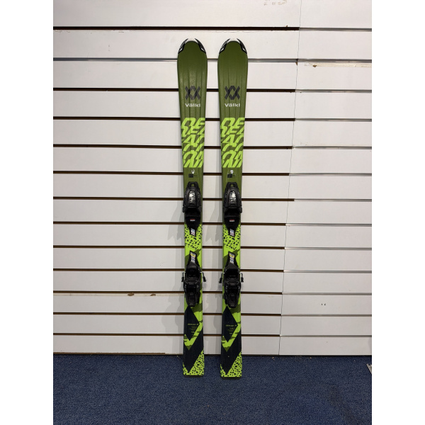 Paire complète de skis enfant Völkl Deacon JR vert olive avec lettres vert citron, logo Völkl et fixations noires, devant un mur à lamelles