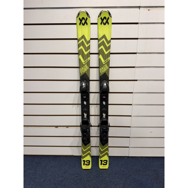 Paire complète de skis Völkl Racetiger JR jaune et noir à motif chevrons, inscription RACE TIGER et fixations noires, devant un mur à lamelles