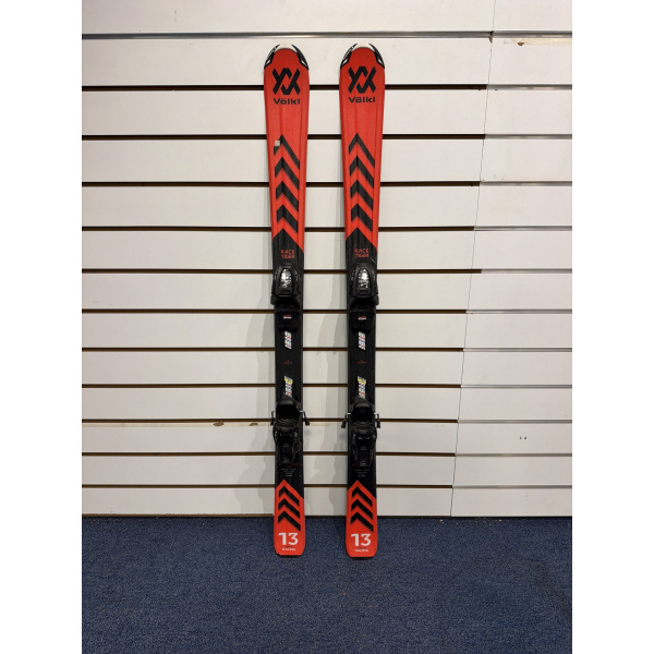 Paire complète de skis Völkl Racetiger JR rouge et noir avec logo Völkl noir et motif chevrons, devant un mur à lamelles