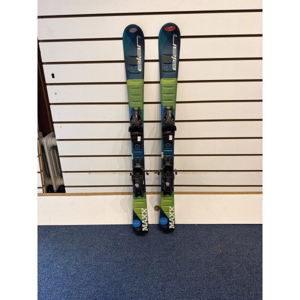 Paire complète de skis junior Elan MAXX Blue à spatules bleu marine, partie basse verte et bleue, fixations noires, devant un mur à lamelles
